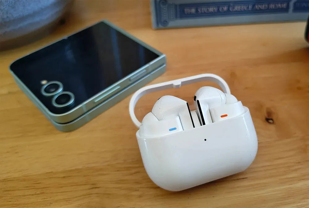White wireless earbuds in a charging case on a wooden surface with a smartphone in the background.