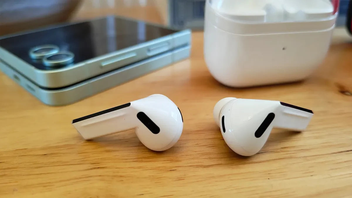White wireless earbuds on a wooden surface with a smartphone and white case in the background.